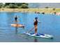 Δύο άτομα κάνουν paddleboarding σε μια λίμνη με πράσινους λόφους στο παρασκήνιο.