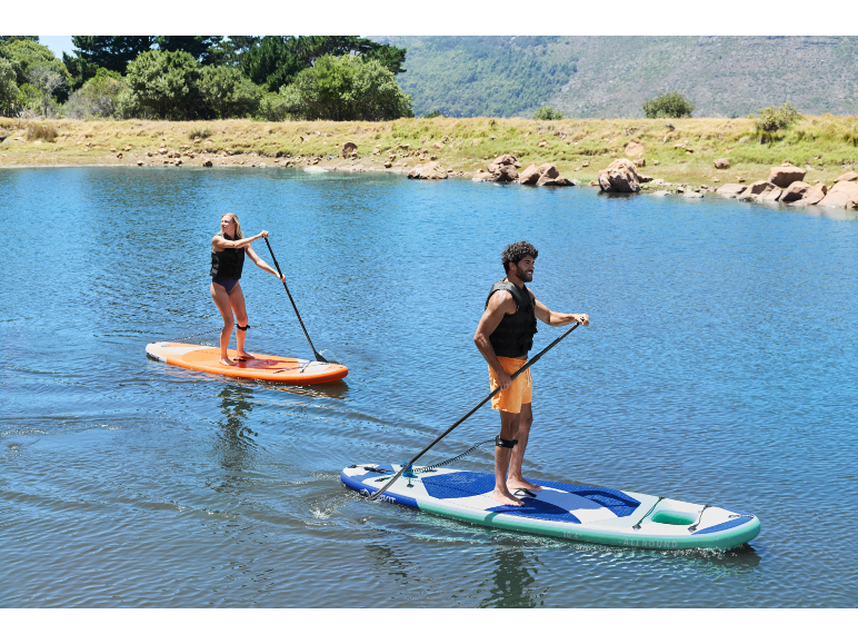 Δύο άτομα κάνουν paddleboarding σε μια λίμνη με πράσινους λόφους στο παρασκήνιο.