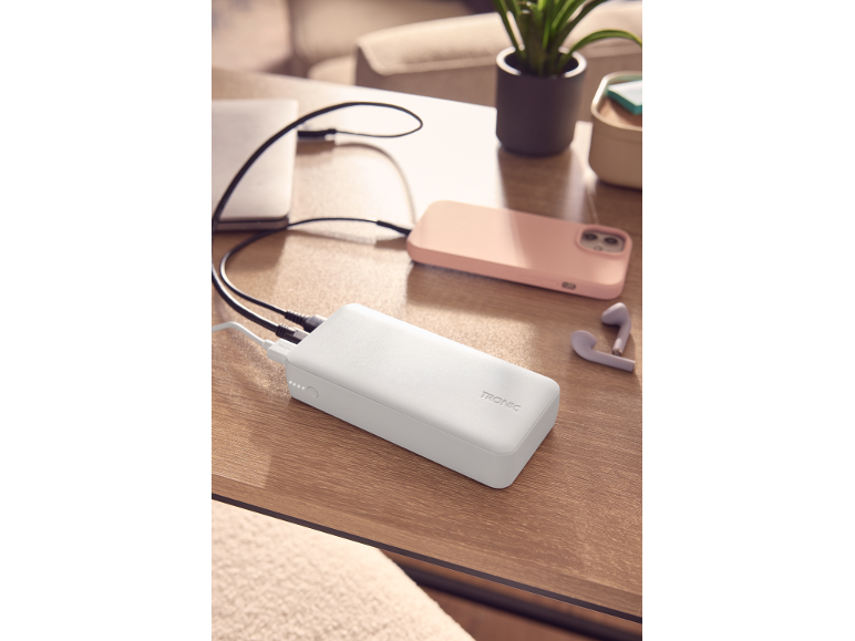 Λευκό power bank φορτίζει ένα ροζ smartphone και ένα laptop σε ξύλινο τραπέζι.