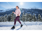 Γυναίκα με ροζ μπουφάν και μωβ κολάν κάνει nordic walking σε χιονισμένο τοπίο.