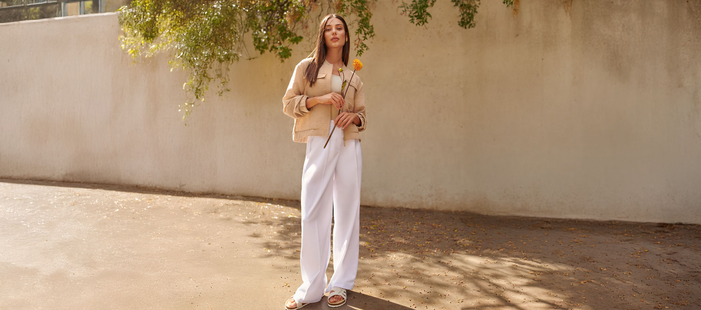 Γυναίκα με μπεζ σακάκι, λευκό παντελόνι και σανδάλια, κρατώντας ένα κίτρινο λουλούδι.