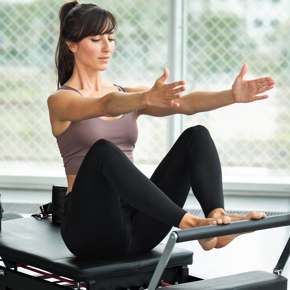 Γυναίκα κάνει Pilates σε reformer, φορώντας τοπ και μαύρο κολάν.