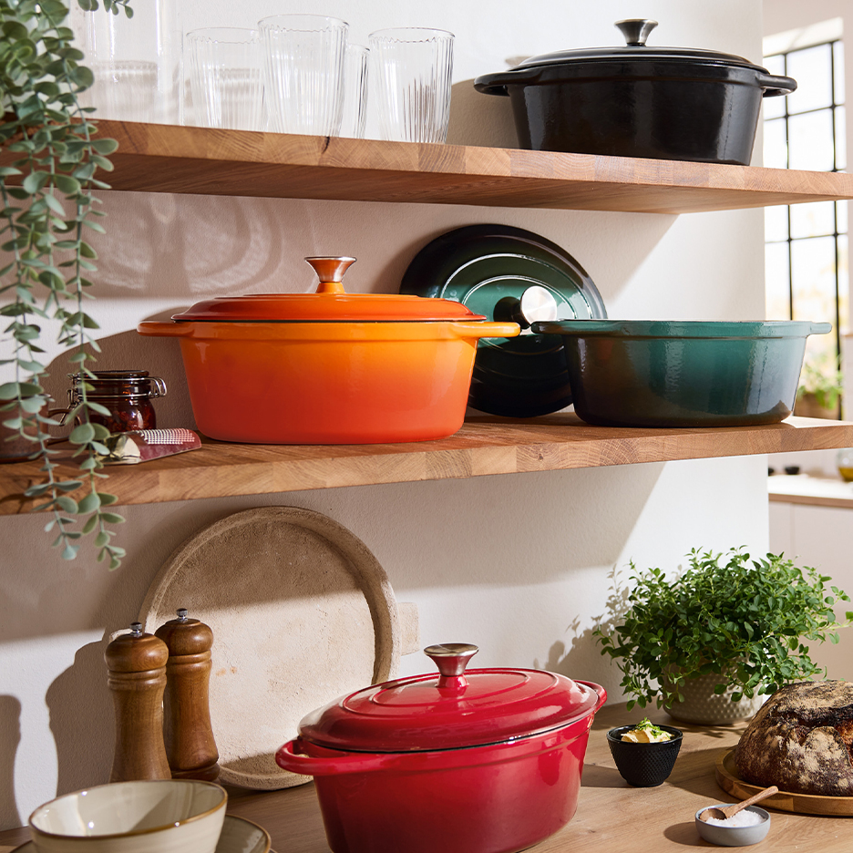 Πολύχρωμες χυτοσιδηρές κατσαρόλες σε ξύλινα ράφια σε μια φωτεινή κουζίνα.