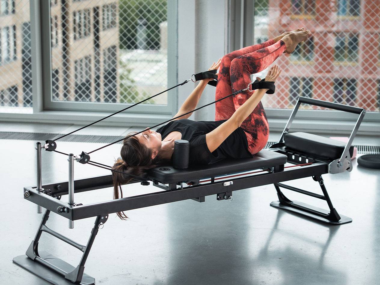 Γυναίκα κάνει Pilates σε reformer με κόκκινο κολάν γιόγκα.