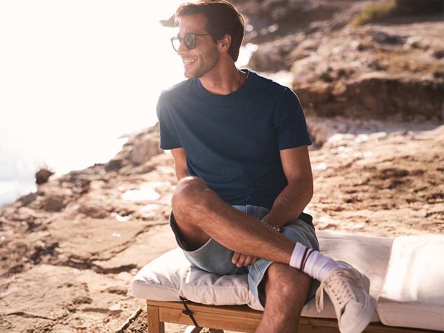 Άνδρας με μπλε t-shirt και λευκά sneakers χαλαρώνει σε ξύλινο παγκάκι δίπλα στη θάλασσα.