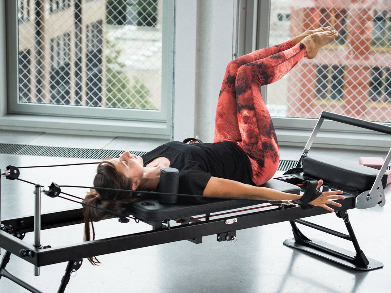Γυναίκα κάνει Pilates σε reformer, με κόκκινο και μαύρο κολάν