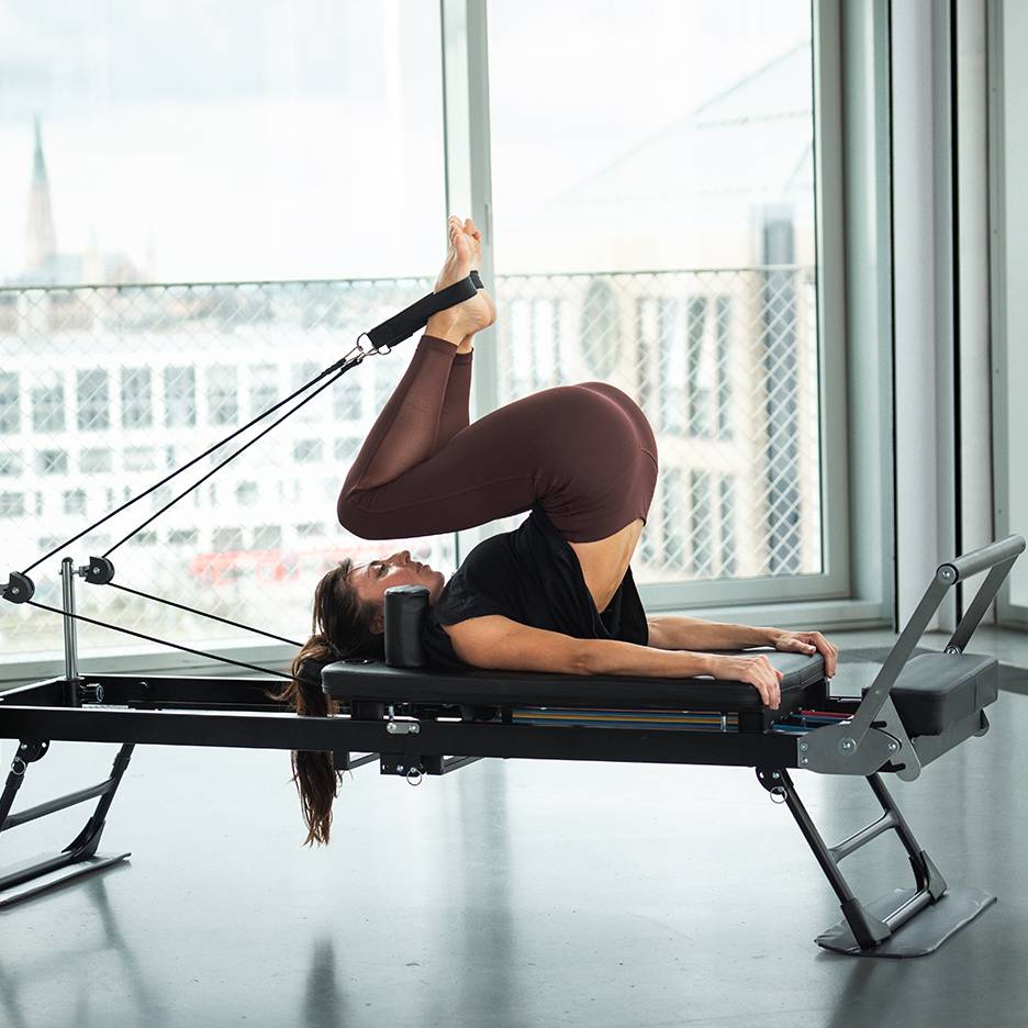Γυναίκα κάνει Pilates σε reformer με τα πόδια ψηλά και δεμένα με ιμάντες.