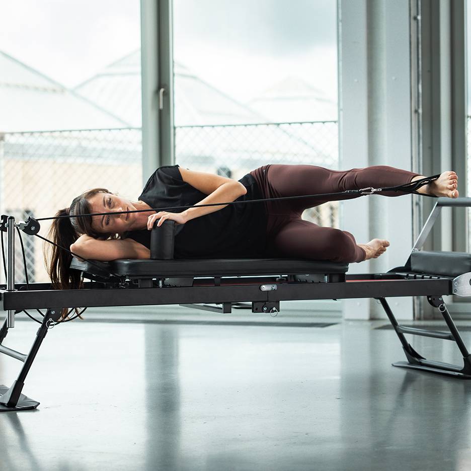 Γυναίκα κάνει Pilates σε μηχάνημα reformer, ξαπλωμένη στο πλάι με ιμάντες ποδιών.