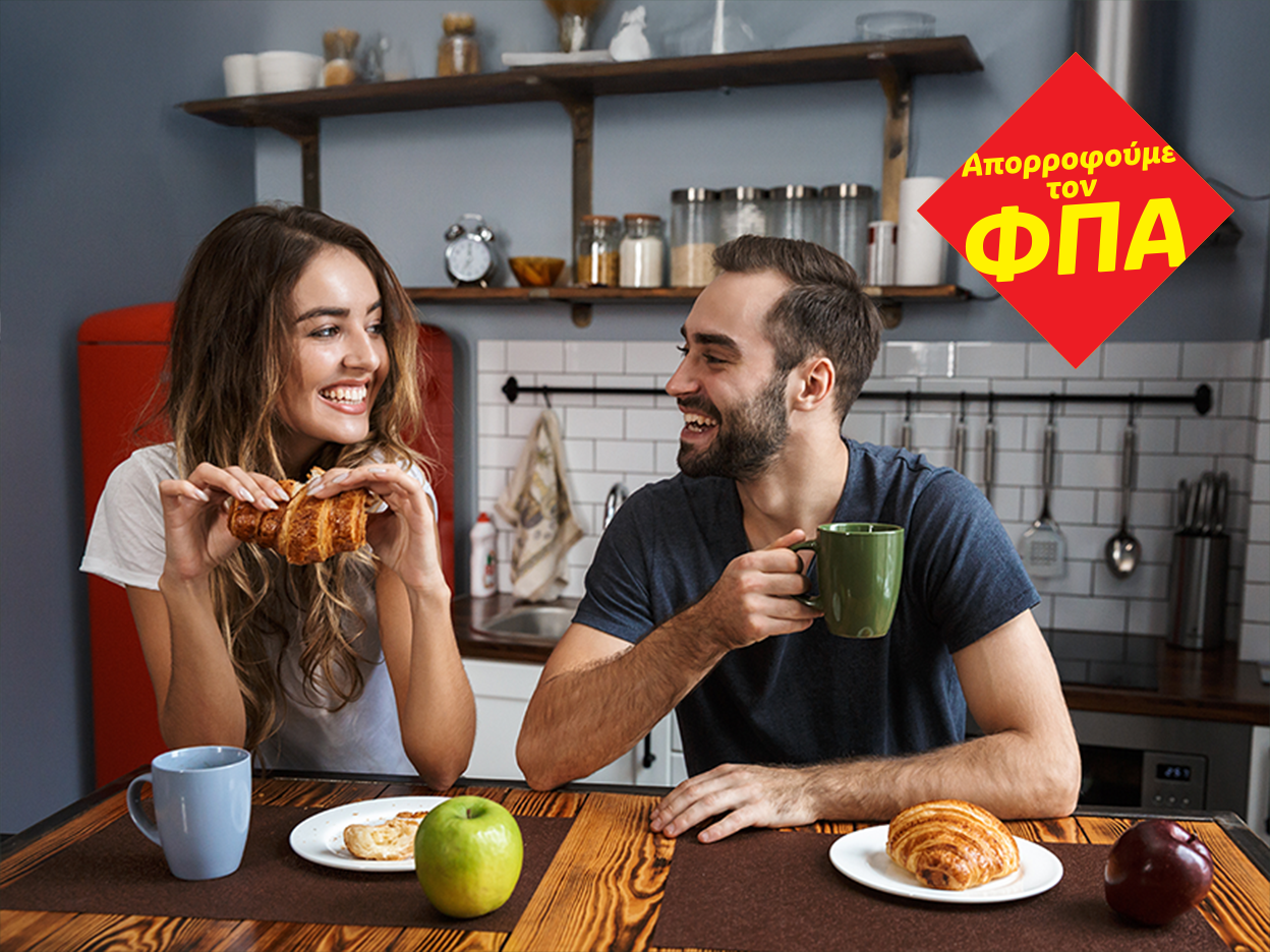 Ζευγάρι απολαμβάνει πρωινό με κρουασάν και καφέ στην κουζίνα, με σήμα «Απορροφούμε τον ΦΠΑ».
