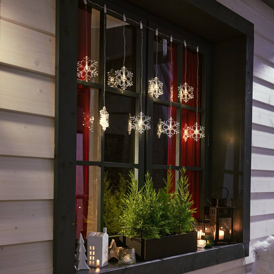 Φωτάκια χιονονιφάδων κρεμασμένα σε παράθυρο με μικρά έλατα και κεριά στο περβάζι.