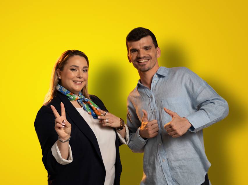 Δύο χαμογελαστοί άνθρωποι, μια γυναίκα και ένας άνδρας, ποζάρουν με χειρονομίες νίκης και μπράβο.