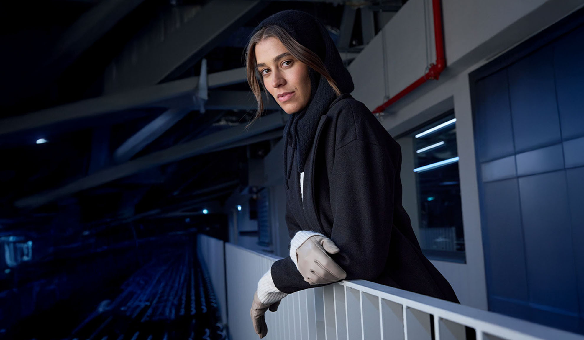 Γυναίκα με μαύρο παλτό και κουκούλα, ακουμπισμένη σε κάγκελα σε μοντέρνο περιβάλλον.