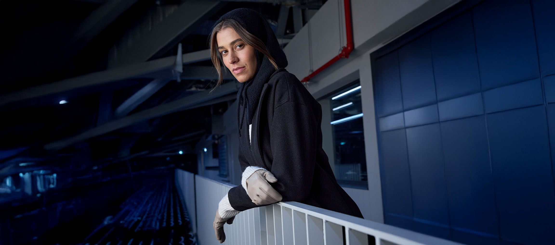 Γυναίκα με μαύρο παλτό και γάντια ακουμπά σε κάγκελα σε αστικό περιβάλλον.