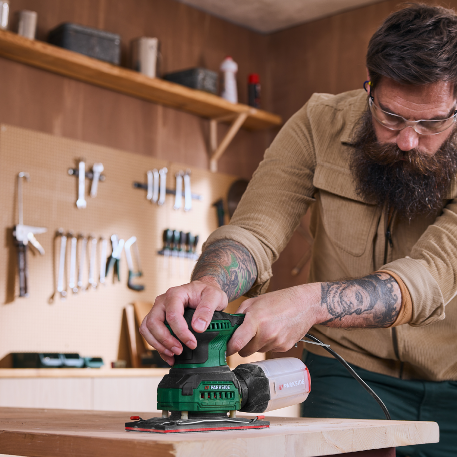 Άνδρας με γενειάδα χρησιμοποιεί τριβείο Parkside σε ξύλινη επιφάνεια σε εργαστήριο.