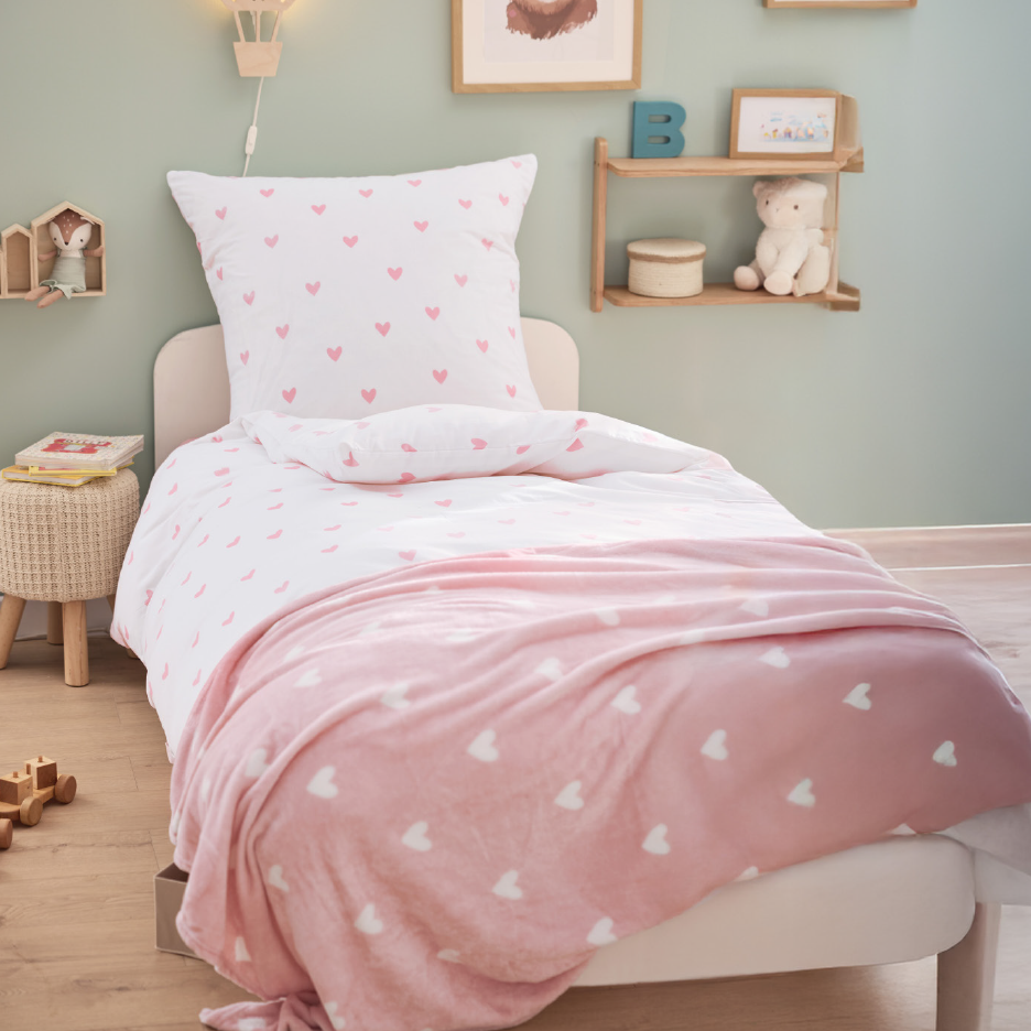 Παιδικό κρεβάτι με λευκά σεντόνια με ροζ καρδιές και ροζ κουβέρτα με λευκές καρδιές.