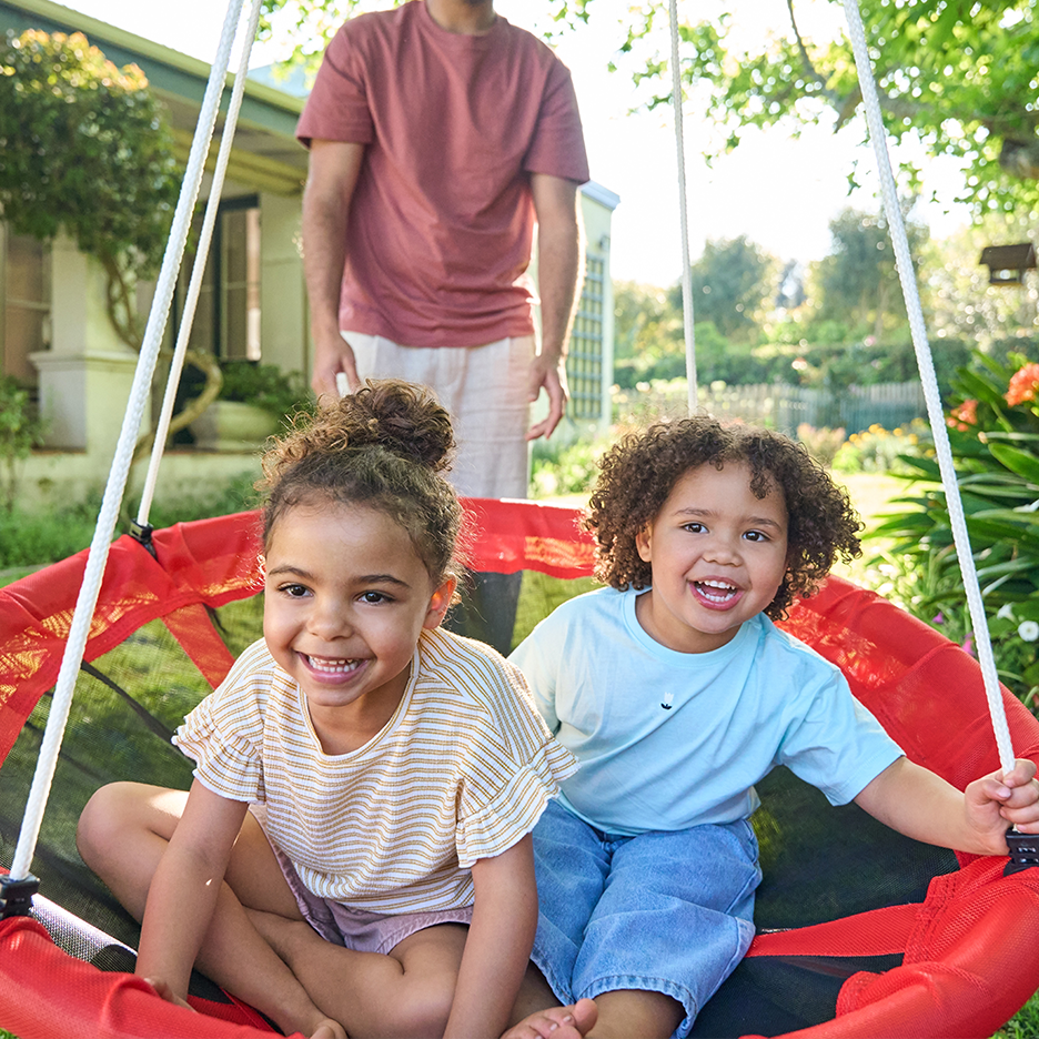 Δύο παιδιά χαμογελούν σε μια κόκκινη κούνια, με έναν ενήλικα να στέκεται από πίσω.