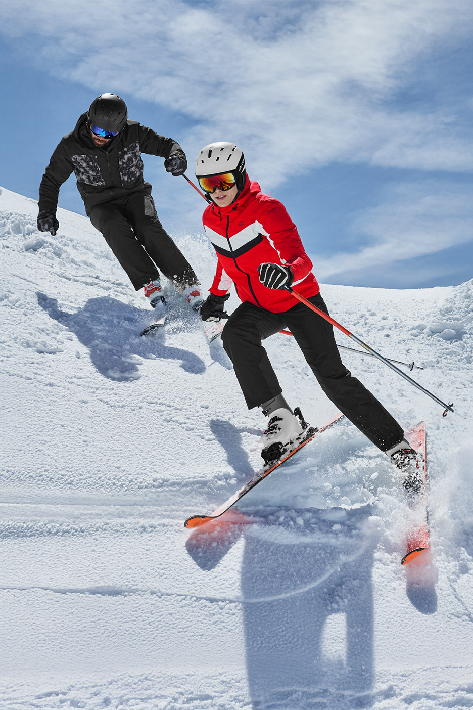 Δύο σκιέρ με μπουφάν σκι, παντελόνια σκι, κράνη και γυαλιά σκι κατεβαίνουν μια χιονισμένη πλαγιά.