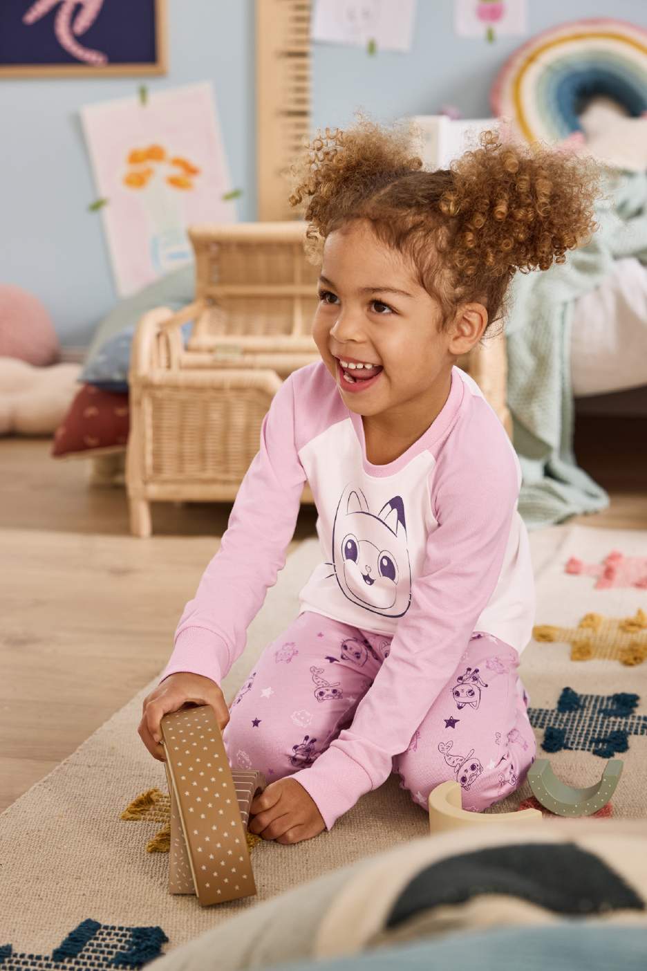 Ένα χαμογελαστό κορίτσι με ροζ πιτζάμες με σχέδιο γάτας παίζει στο πάτωμα.