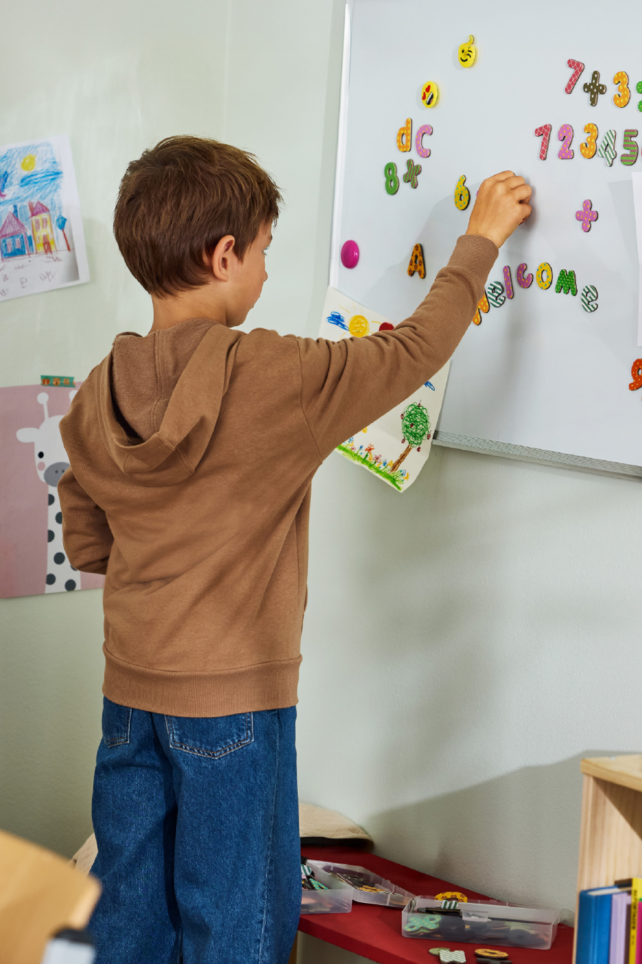 Παιδί με φούτερ και τζιν παίζει με μαγνητικά γράμματα και αριθμούς σε λευκό πίνακα.