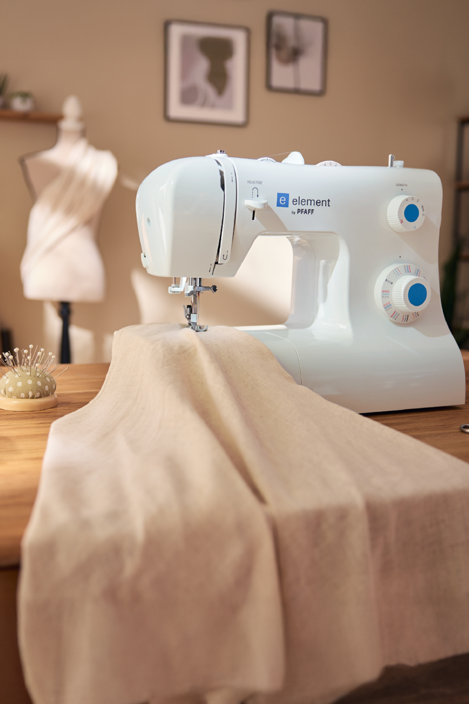 Λευκή ραπτομηχανή Pfaff με ύφασμα σε ξύλινο τραπέζι, με μανεκέν στο παρασκήνιο.