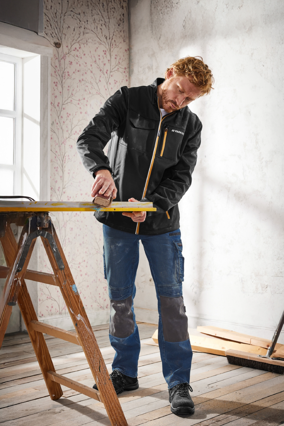 Άνδρας με μπουφάν και παντελόνι εργασίας Parkside τρίβει ξύλο σε ένα δωμάτιο.
