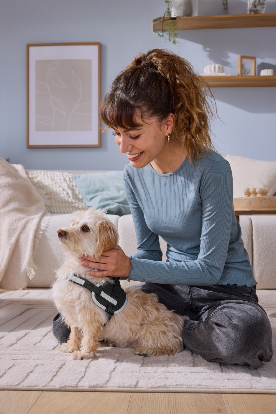 Γυναίκα χαμογελάει και χαϊδεύει ένα μικρό σκυλί με λουρί, καθισμένη σε χαλί.