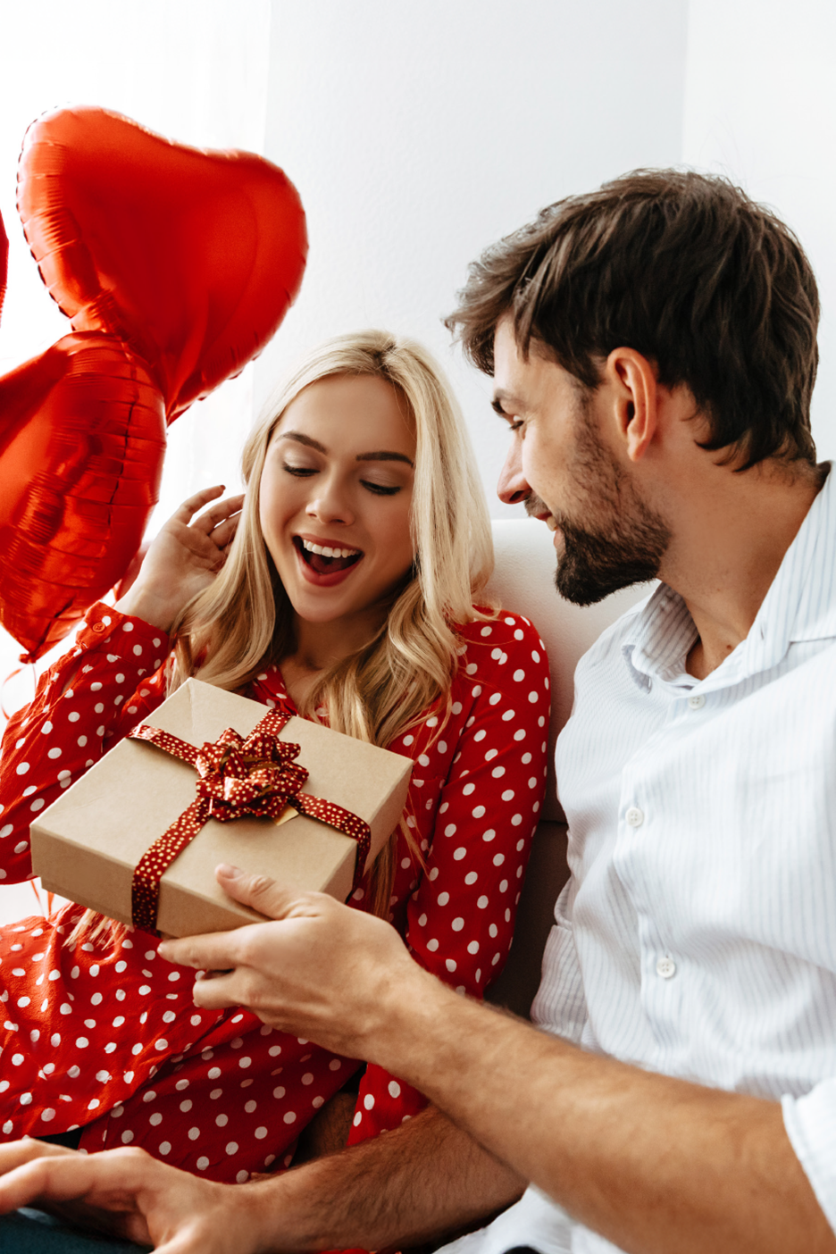 Ένας άντρας δίνει ένα δώρο σε μια χαρούμενη γυναίκα με κόκκινα μπαλόνια σε σχήμα καρδιάς.
