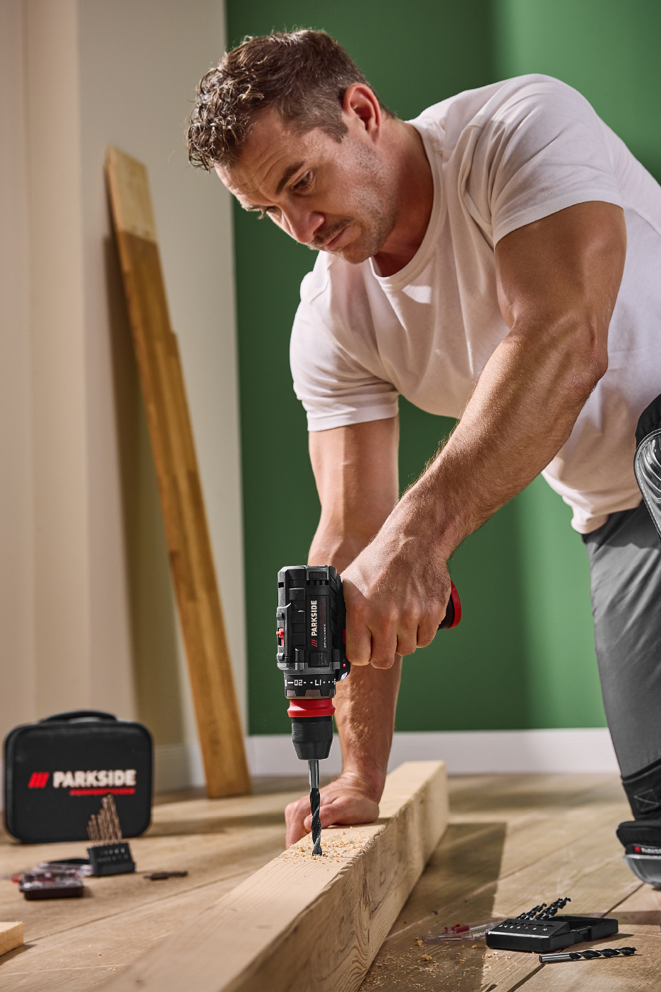 Άντρας χρησιμοποιεί ένα ασύρματο δράπανο για να τρυπήσει μια ξύλινη σανίδα, με ένα σετ τρυπανιών στο πάτωμα.