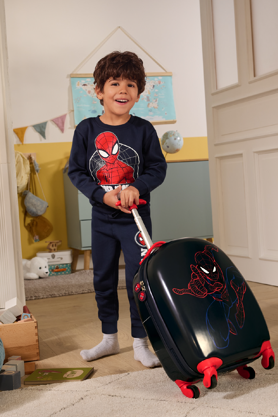 Χαμογελαστό αγόρι με μπλε φόρμα με σχέδιο υπερήρωα, σέρνοντας μια βαλίτσα με τον ίδιο υπερήρωα.