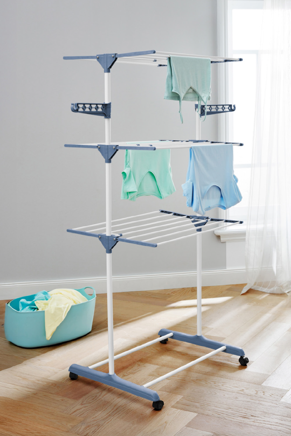 Τριώροφη απλώστρα ρούχων με ρόδες, με μερικά ρούχα απλωμένα και ένα καλάθι με ρούχα στο πάτωμα.