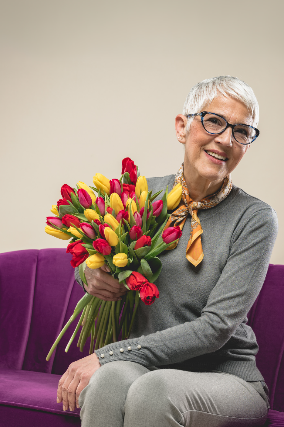 Χαμογελαστή ηλικιωμένη γυναίκα με γυαλιά, κρατώντας μια μεγάλη ανθοδέσμη με κόκκινες και κίτρινες τουλίπες.