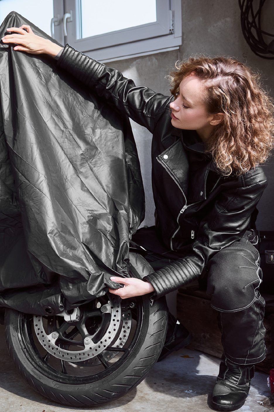Γυναίκα με δερμάτινο μπουφάν και παντελόνι καλύπτει μοτοσικλέτα με μαύρο κάλυμμα.