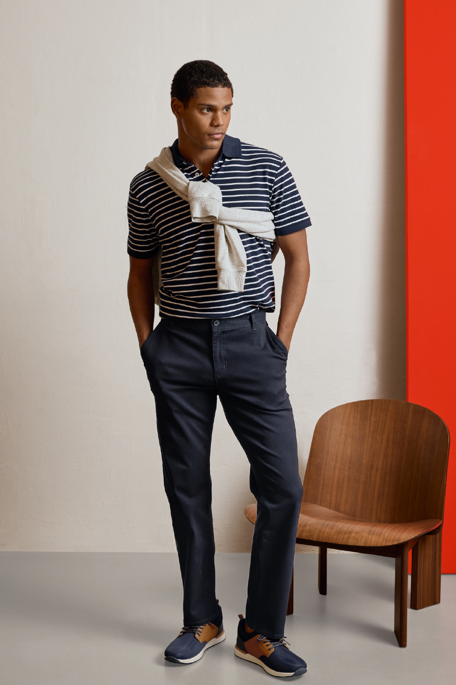 Άνδρας με ριγέ μπλουζάκι πόλο, σκούρο μπλε παντελόνι και γκρι πουλόβερ στους ώμους.