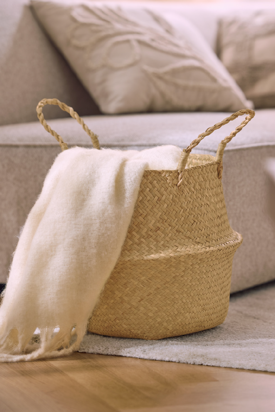 Ψάθινο καλάθι με λευκή κουβέρτα δίπλα σε έναν καναπέ με μαξιλάρια.