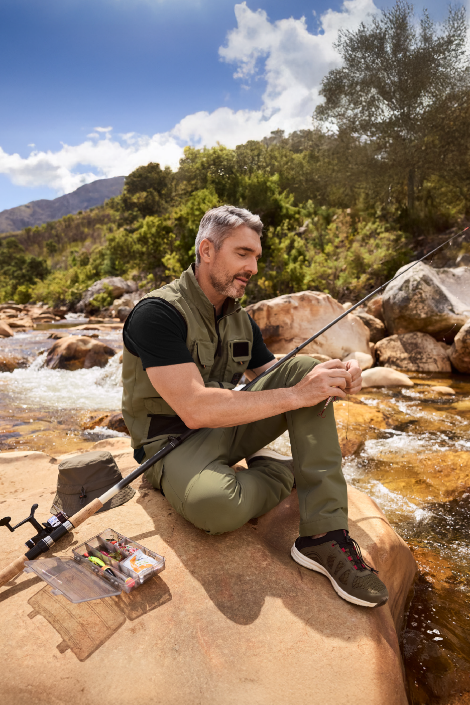 Άνδρας με χακί γιλέκο και παντελόνι ψαρεύει σε ποτάμι με βουνά στο βάθος.