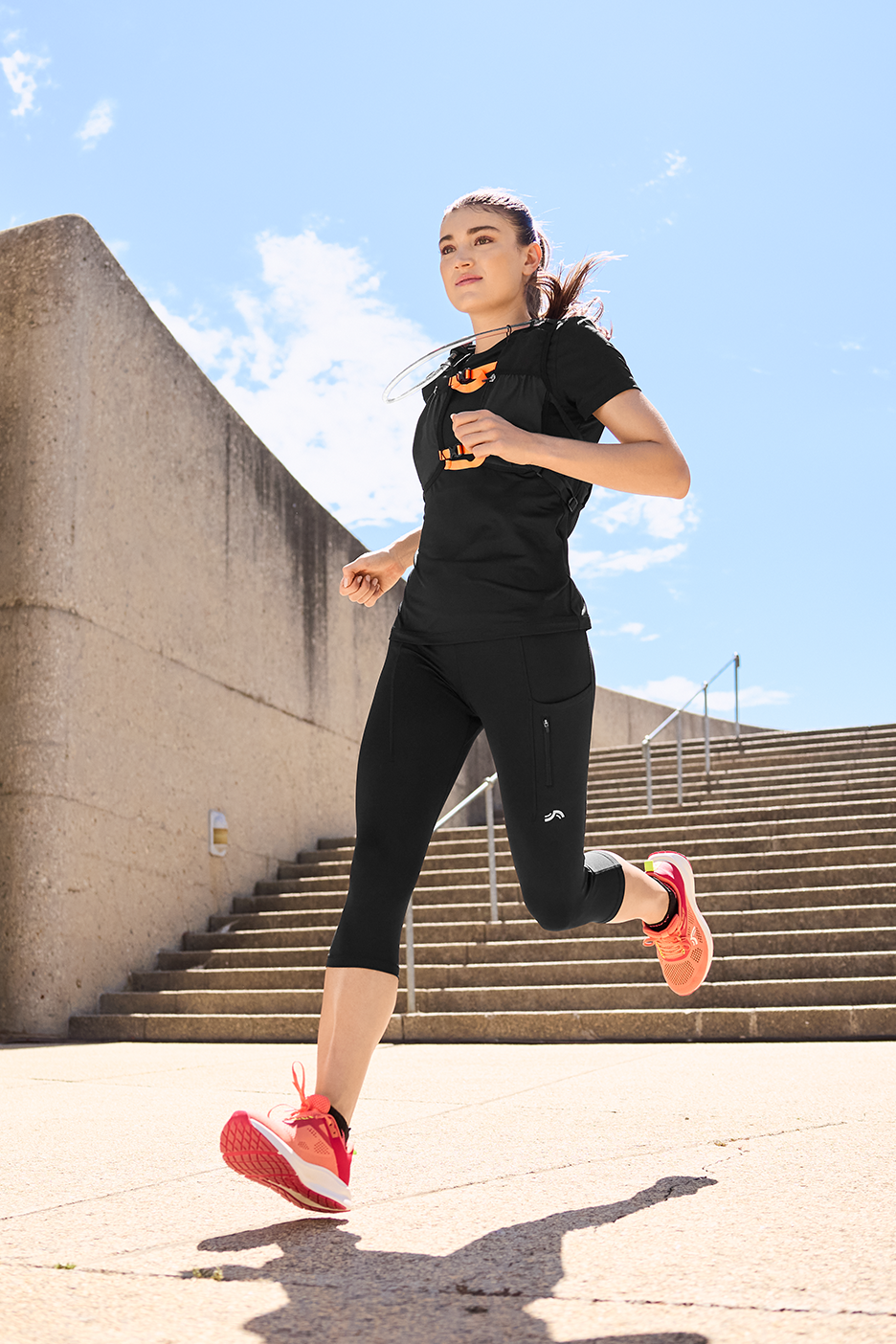 Γυναίκα με μαύρο αθλητικό μπλουζάκι, κολάν και πορτοκαλί αθλητικά παπούτσια τρέχει σε εξωτερικό χώρο.