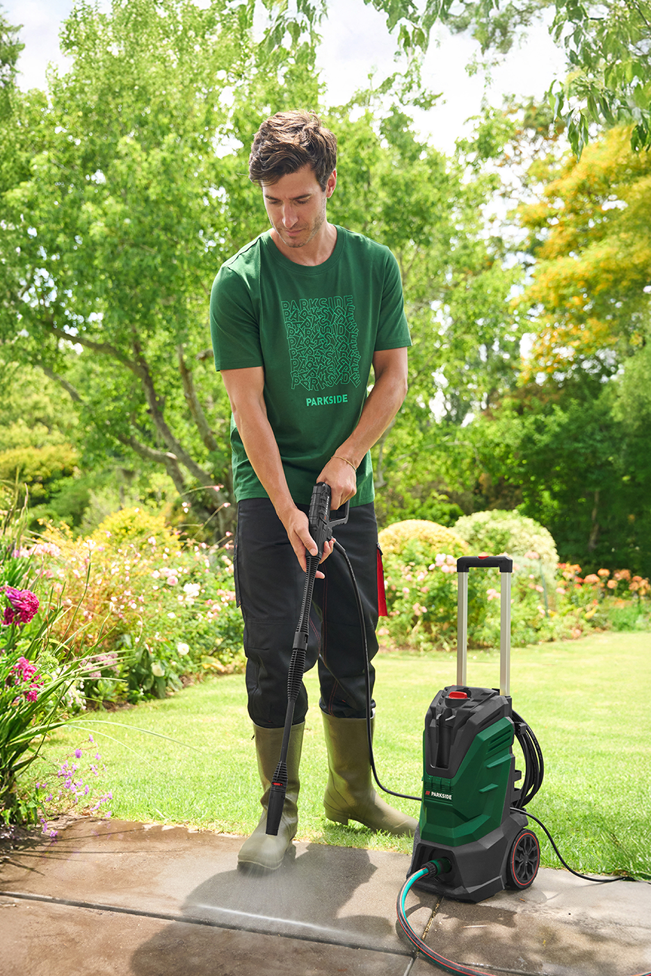 Άντρας καθαρίζει ένα μονοπάτι με ένα πλυστικό μηχάνημα υψηλής πίεσης σε έναν καταπράσινο κήπο.