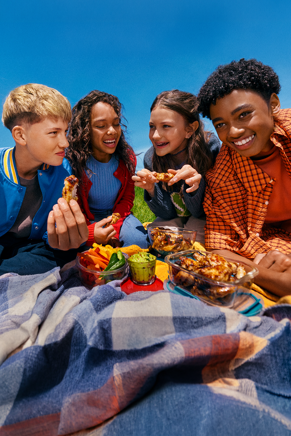 Τέσσερα παιδιά κάνουν πικνίκ σε εξωτερικό χώρο, τρώνε φτερούγες κοτόπουλου, λαχανικά και γουακαμόλε.