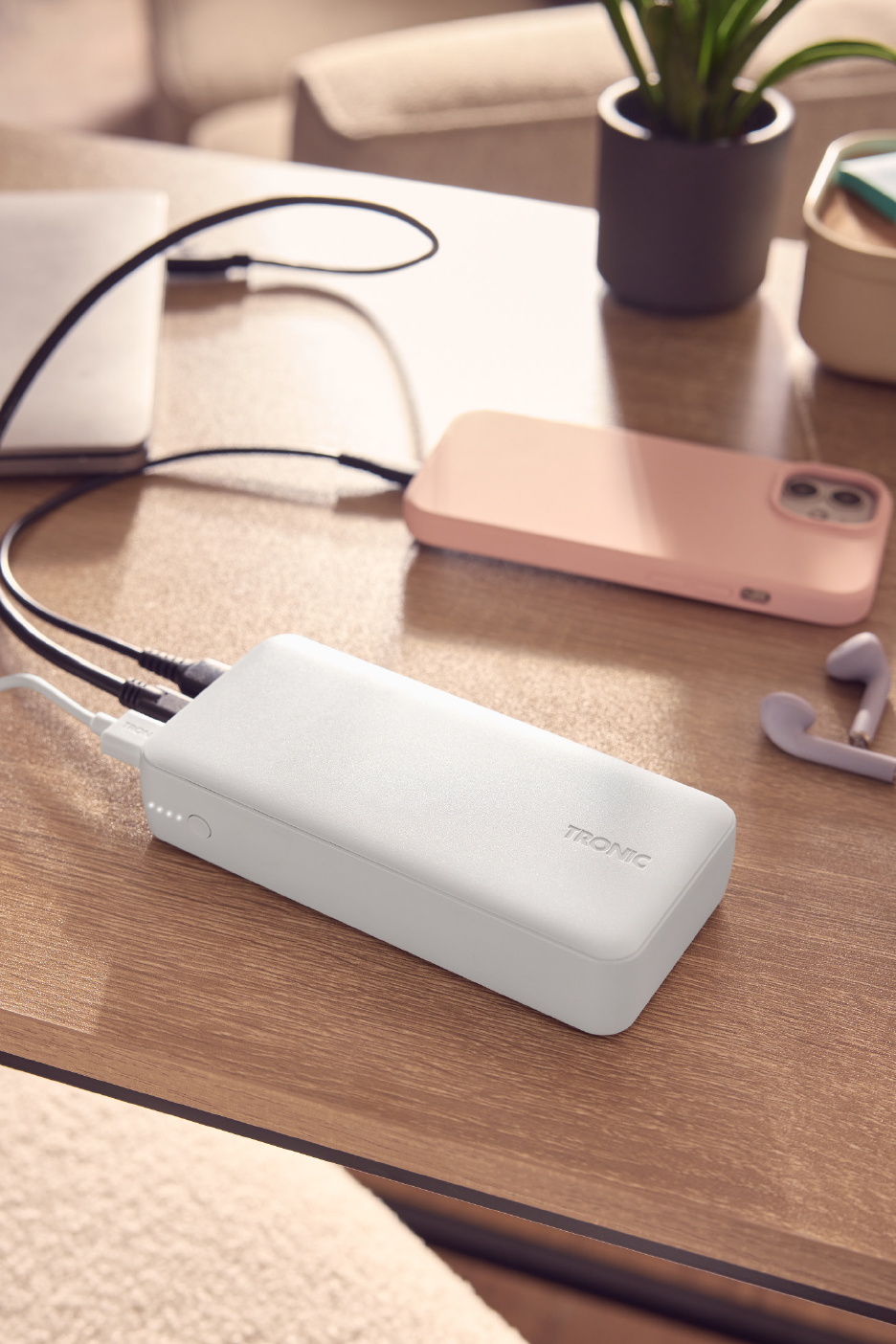 Λευκό power bank φορτίζει ένα ροζ smartphone και ένα laptop σε ένα ξύλινο τραπέζι.