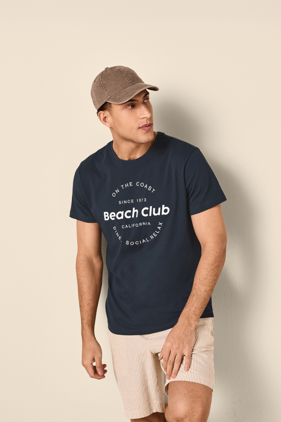 Άνδρας με σκούρο μπλε μπλουζάκι με στάμπα 'Beach Club' και ριγέ σορτς.