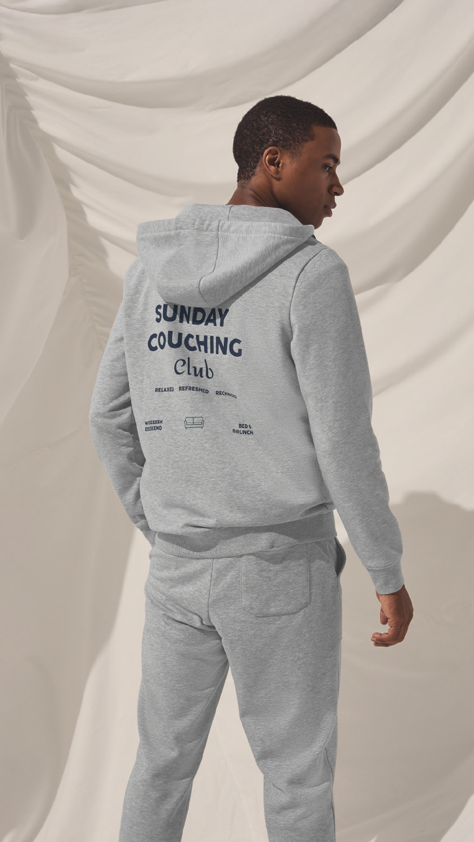 Άνδρας με γκρι φούτερ και παντελόνι φόρμας 'Sunday Couching Club' με σχέδιο καναπέ.