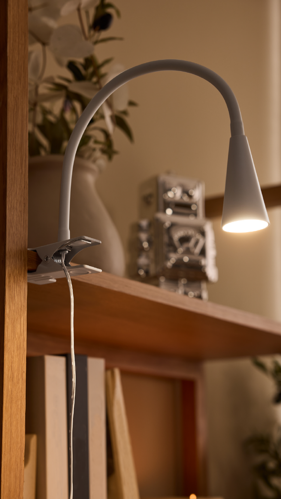 Λευκό φωτιστικό γραφείου με κλιπ σε ξύλινο ράφι, με διακοσμητικά στο βάθος.