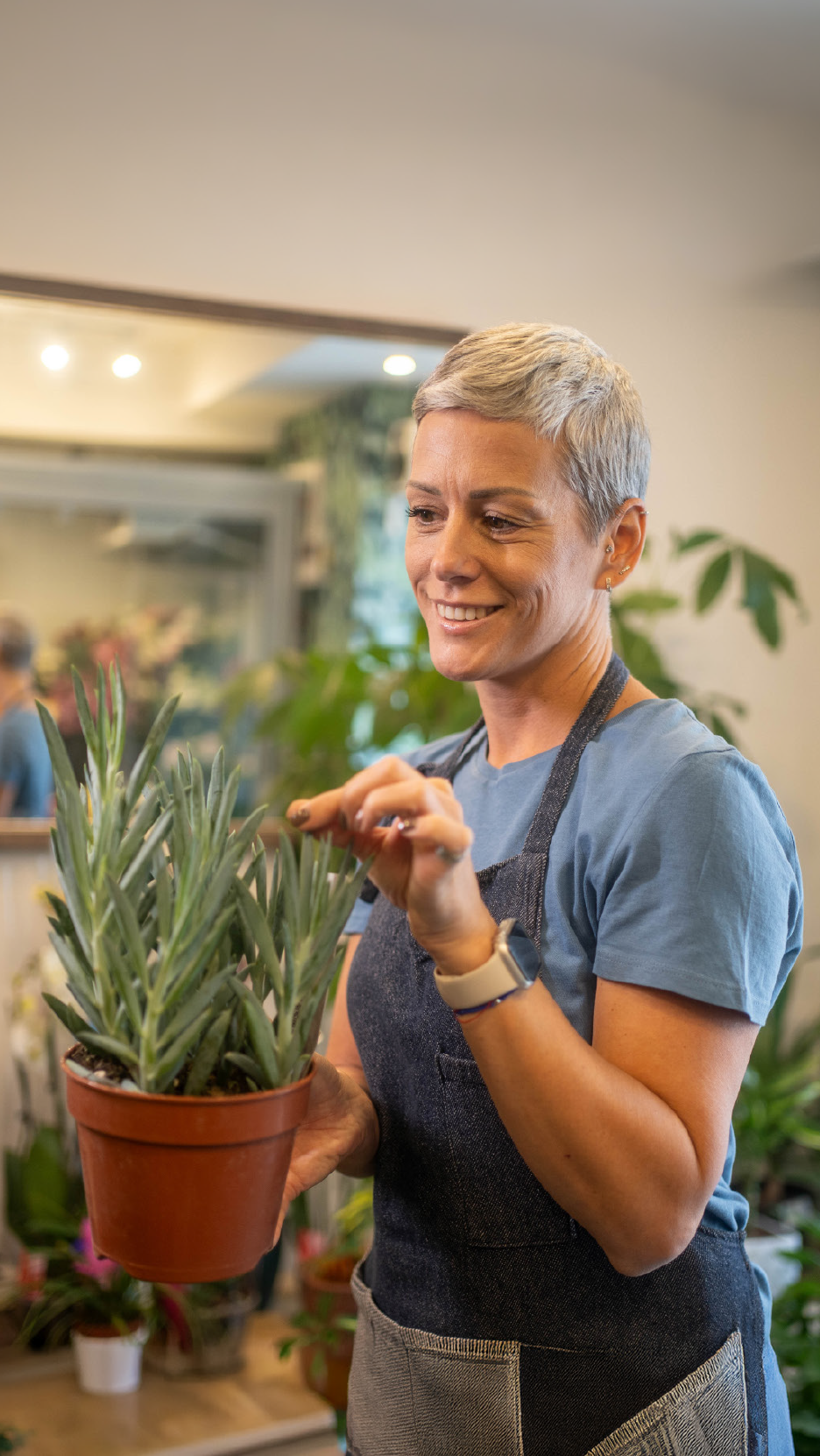 Μια χαμογελαστή γυναίκα με ποδιά φροντίζει ένα φυτό σε γλάστρα σε ένα κατάστημα.