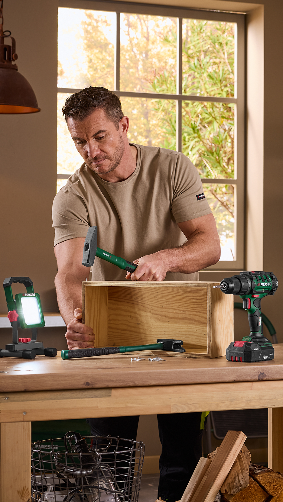 Άντρας συναρμολογεί ένα ξύλινο κουτί με σφυρί, δίπλα του ένα τρυπάνι και ένας προβολέας.