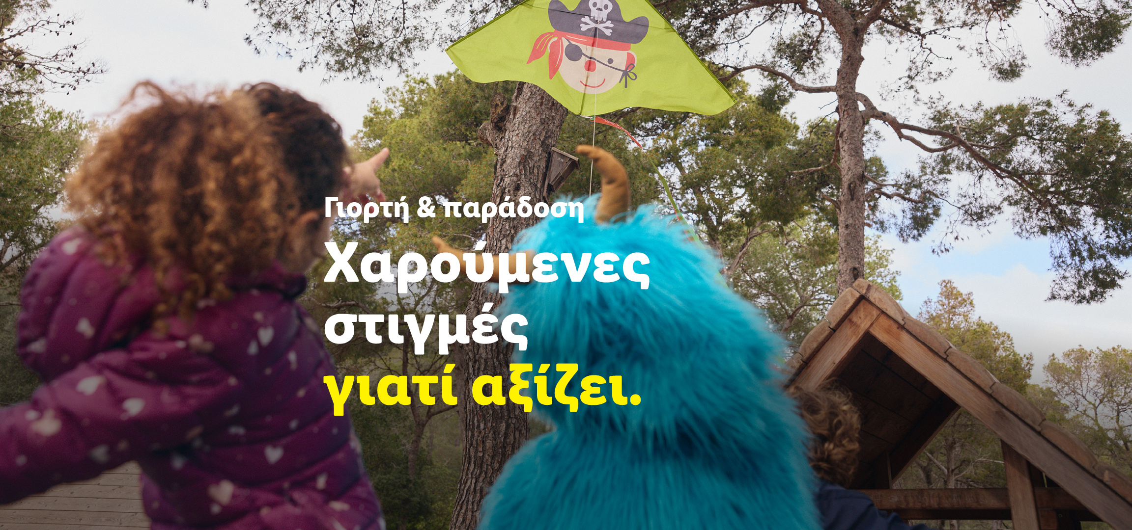 Παιδιά παίζουν με χαρταετό σε δάσος, με κείμενο: «Γιορτή & παράδοση. Χαρούμενες στιγμές γιατί αξίζει».