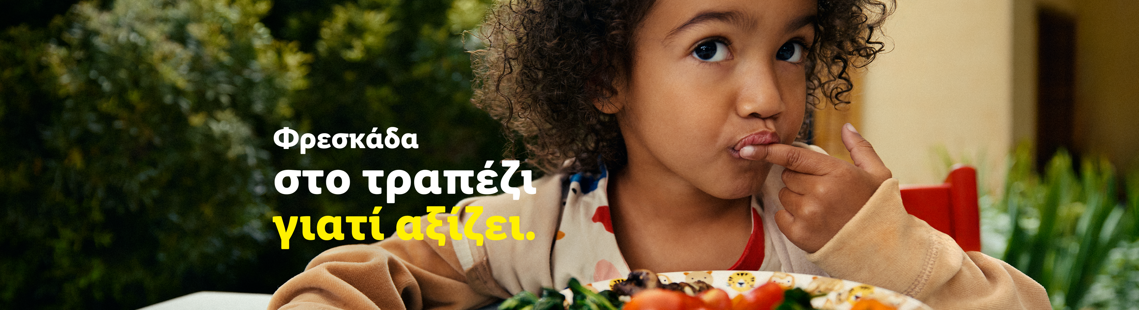 Παιδί με σγουρά μαλλιά τρώει από πιάτο με φαγητό, με κείμενο «Φρεσκάδα στο τραπέζι γιατί αξίζει».