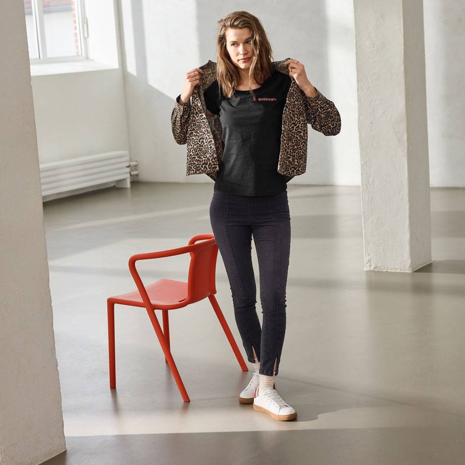 Γυναίκα με μαύρο μπλουζάκι με επιγραφή, τζιν και λεοπάρ ζακέτα, στέκεται δίπλα σε κόκκινη καρέκλα.