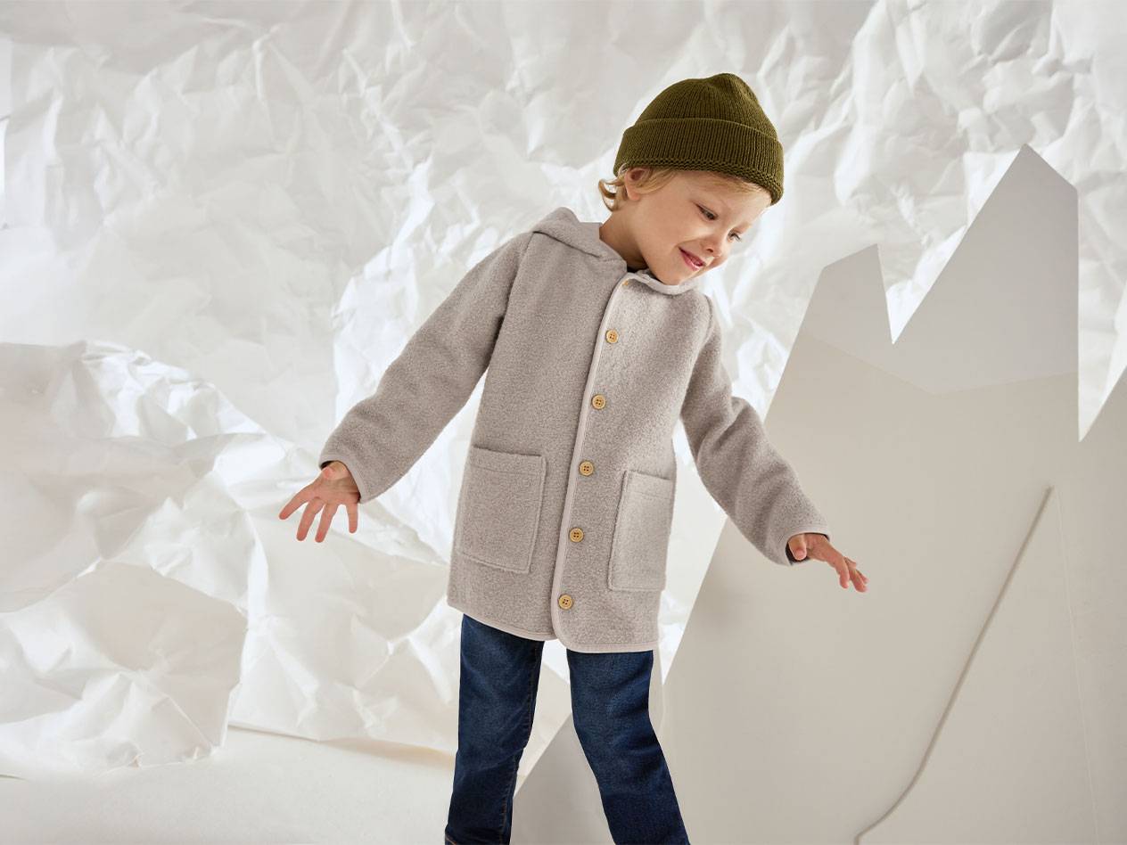 Αγόρι με γκρι μάλλινο μπουφάν και πράσινο σκούφο, χαμογελαστό σε φόντο τσαλακωμένου χαρτιού.