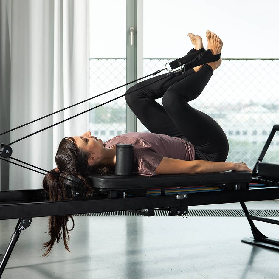 Γυναίκα ασκεί τα πόδια της σε reformer Pilates με λάστιχα αντίστασης.