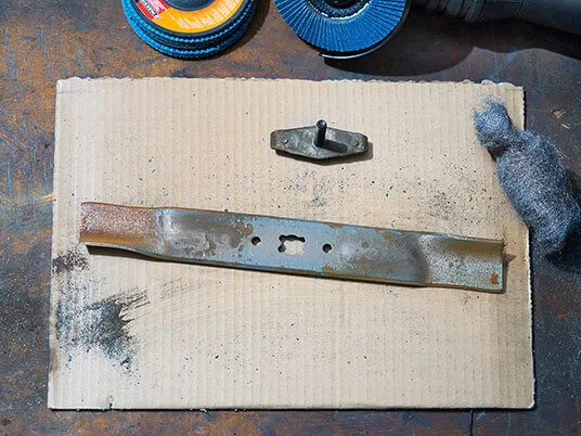 Σκουριασμένη λεπίδα χλοοκοπτικής μηχανής σε χαρτόνι με λειαντικούς δίσκους και ατσαλόμαλλο.